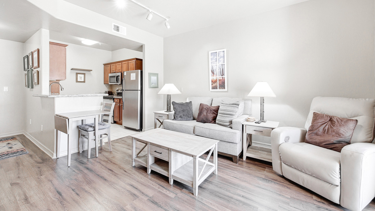 Bright, modern living room with wooden floors, a gray sofa, white armchair, coffee table, two side tables with lamps, and an open kitchen with stainless steel appliances in the background.