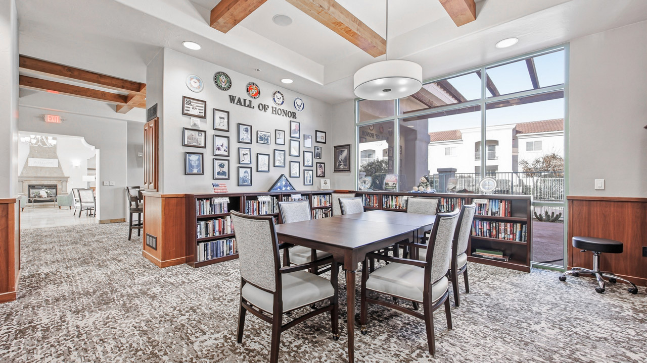 A bright room with a table and four chairs, bookshelves, large windows, and a “Wall of Honor” displaying military insignias, photos, and plaques. Exposed wooden beams line the ceiling, and a modern light fixture hangs above.
