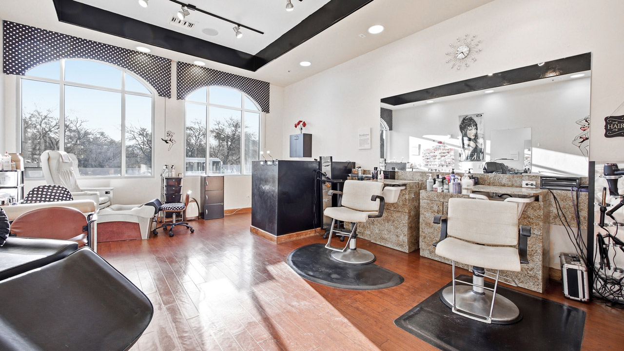 A bright, modern hair salon with large windows, two styling chairs facing a mirror, hair products on counters, and wood floors. Sunlight streams in, highlighting the clean and inviting space.