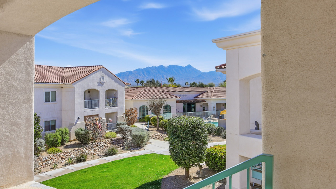 View of a courtyard with green grass, shrubs, and pathways, surrounded by light-colored, stucco apartment buildings. Mountains and a clear blue sky are visible in the distance.