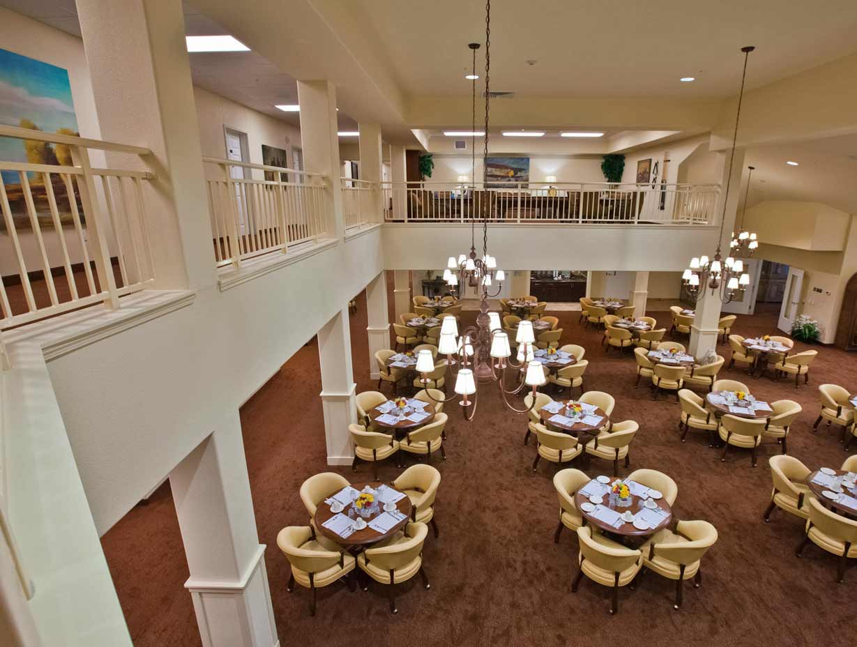 A spacious dining area with two levels featuring neatly arranged tables with beige chairs, white tablecloths, and folded napkins. Chandeliers hang from the ceiling, and there's a brown carpet on the floor. A white railing spans the upper level, which overlooks the space.