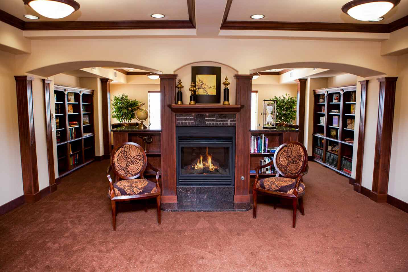 A cozy reading room with a fireplace at the center, flanked by two cushioned chairs with wooden frames. Bookshelves filled with books and decorative items line the walls on either side. The warm, inviting space features carpeted floors and soft overhead lighting.