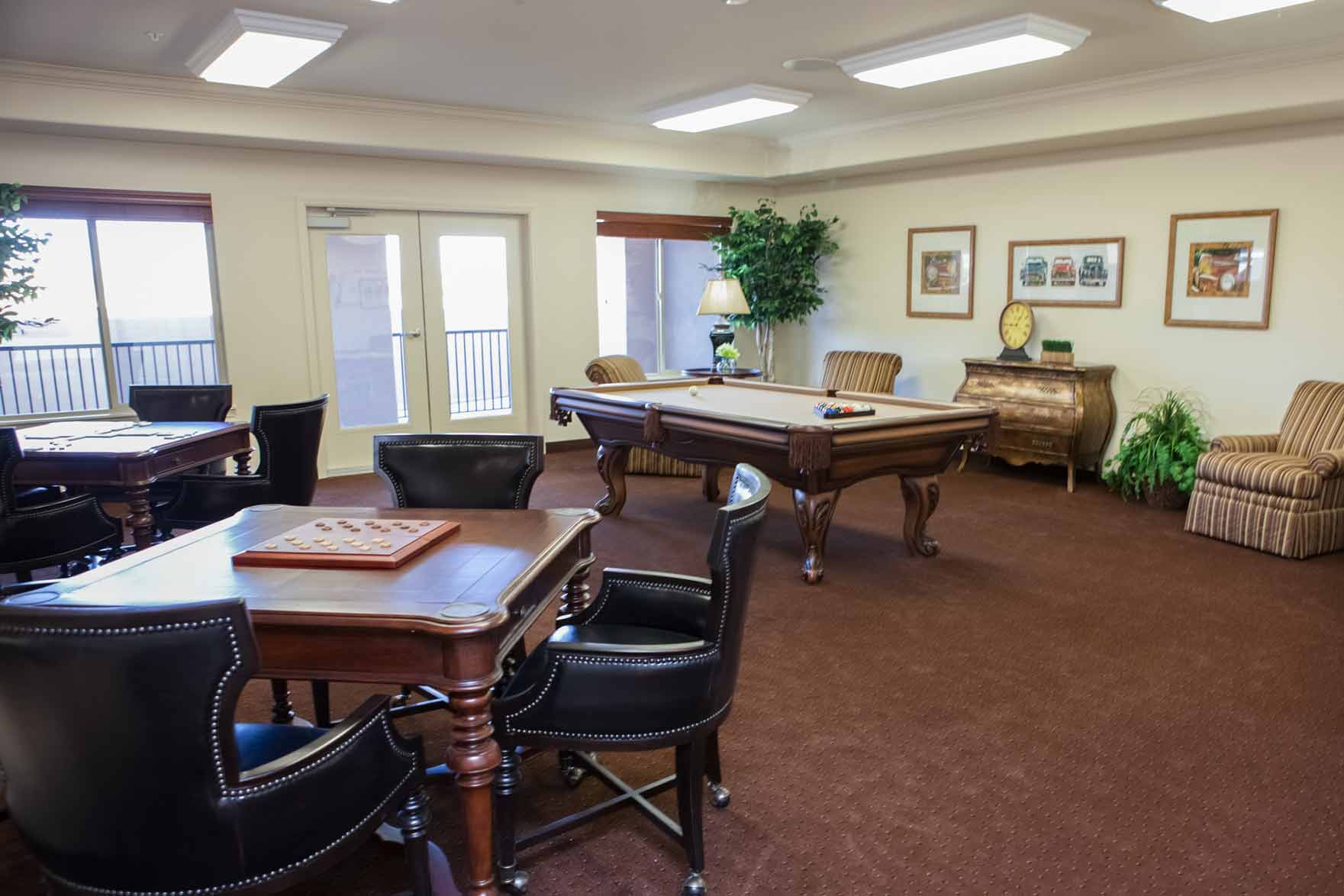 A recreation room with a pool table in the center, surrounded by chairs, lounge chairs, and indoor plants. A game table featuring a checkers set is in the foreground. Sunlight streams through the large windows and glass door, illuminating the space.