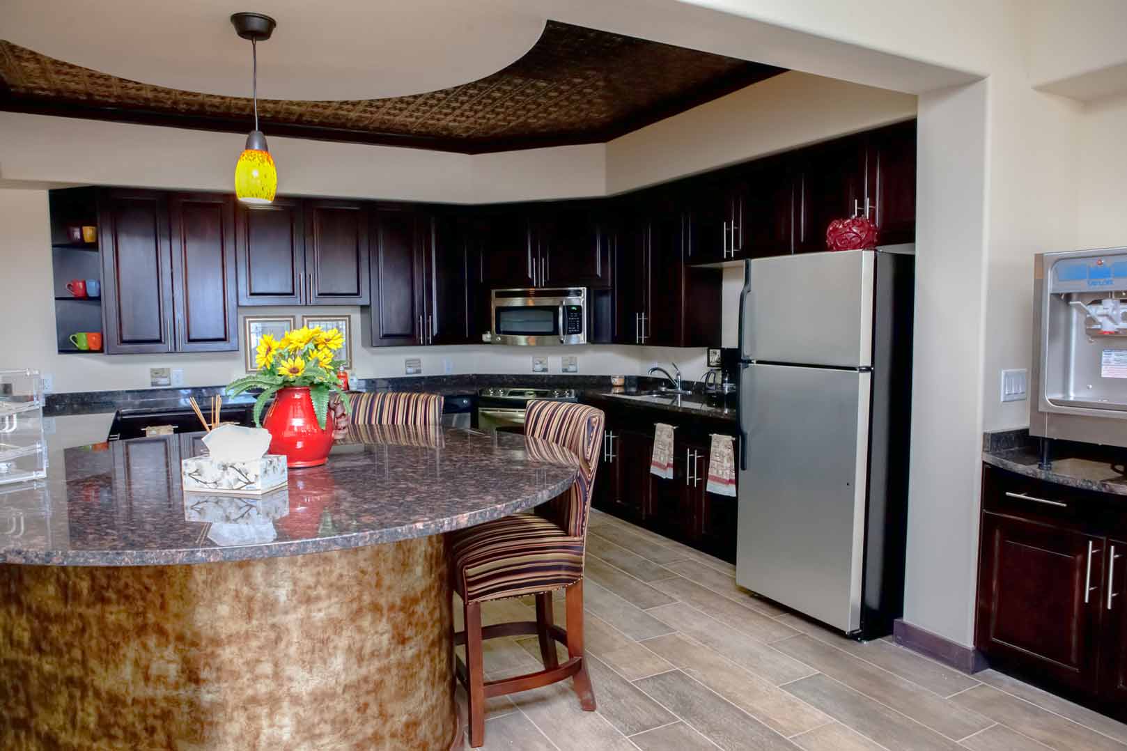 A modern kitchen featuring dark wooden cabinets, stainless steel appliances, a large granite countertop island with two cushioned chairs, and a vase of yellow flowers. Overhead, a yellow pendant light illuminates the island, and there is a textured ceiling detail.