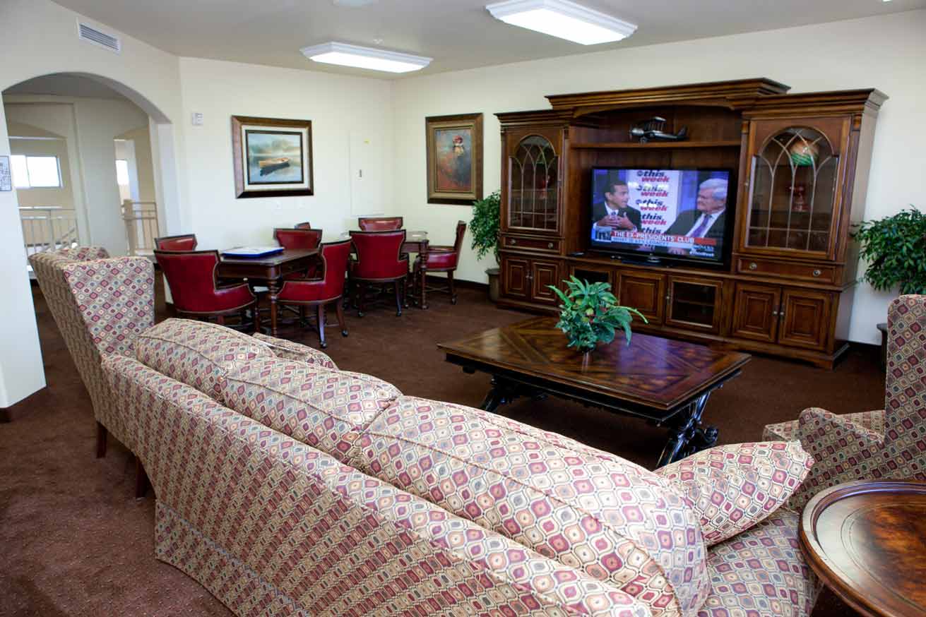 A cozy living room with patterned sofas, a large wooden entertainment center, a flat-screen TV showing a news program, and a dining table with red chairs. The room features warm lighting, framed artwork on the walls, and potted plants in the corners.