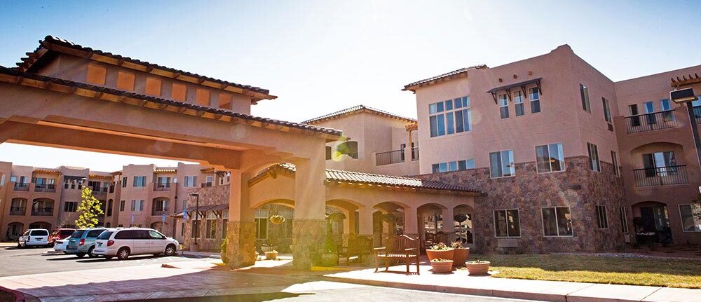 A residential complex featuring stucco buildings with balconies and large windows. The foreground shows a covered driveway with parked cars nearby. The architecture includes elements of Spanish or Mediterranean style, with stone accents and terracotta roof tiles.