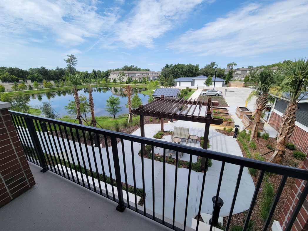 View from a balcony overlooking a serene courtyard featuring a wooden pergola, outdoor seating, and landscaping with palm trees and plants. Beyond the courtyard is a tranquil pond surrounded by greenery and trees under a partly cloudy blue sky.