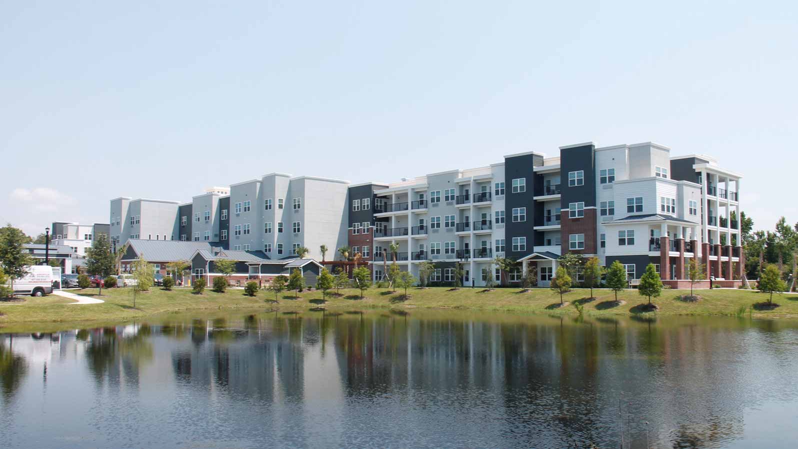 A waterfront view of modern apartment buildings with a mix of gray, white, and black exteriors. The buildings are reflected in the calm water of a pond, surrounded by green lawns and small trees under a clear blue sky.