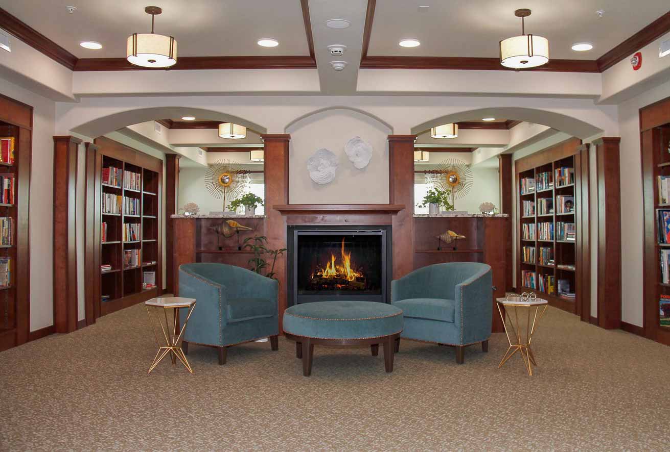 A cozy library lounge featuring two teal armchairs and a matching ottoman centered around a modern fireplace. Flanked by wooden bookshelves filled with books, the room is well-lit with multiple ceiling lights and decorated with vases and artwork.