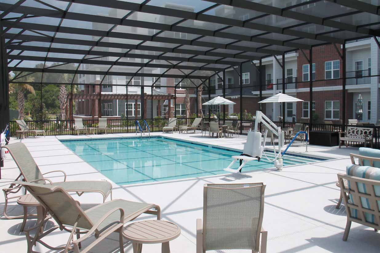 Enclosed outdoor swimming pool area with lounge chairs, tables, and umbrellas. A pool lift chair is installed beside the pool, and there are apartment buildings visible in the background. The area is covered with a metal frame and netting structure.