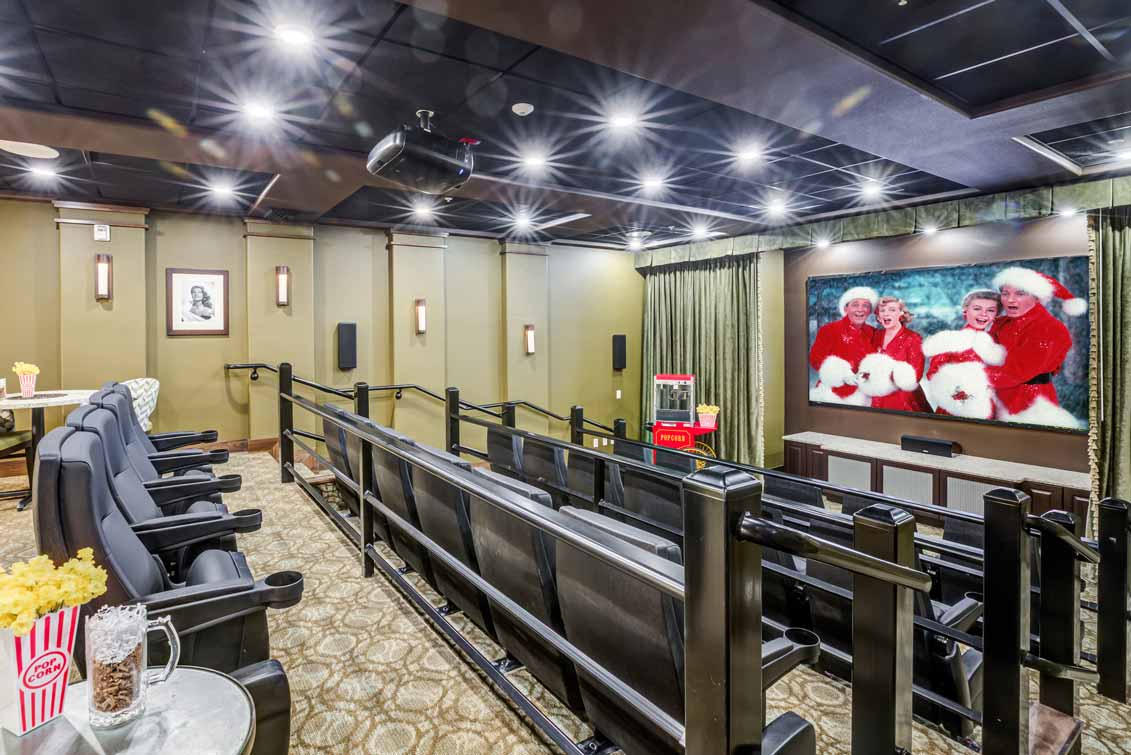 A small private theater with several rows of black seats, showing a movie on a large screen featuring four people dressed as Santa Claus. A popcorn machine stands near the screen, and a bowl of popcorn is on a table in the foreground.