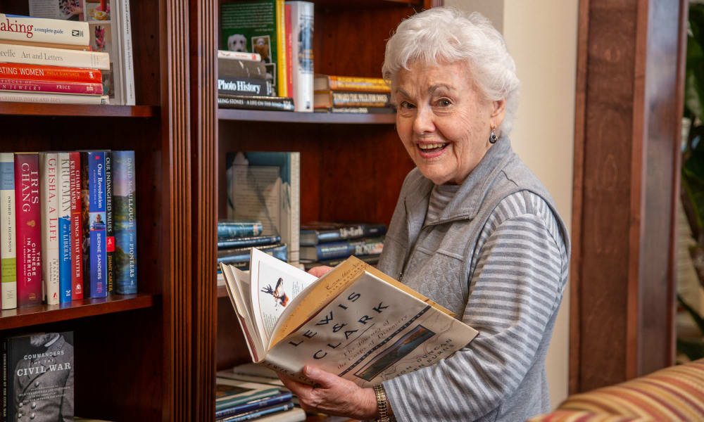 An elderly woman with curly white hair and a striped gray sweater smiles while holding an open book titled 
