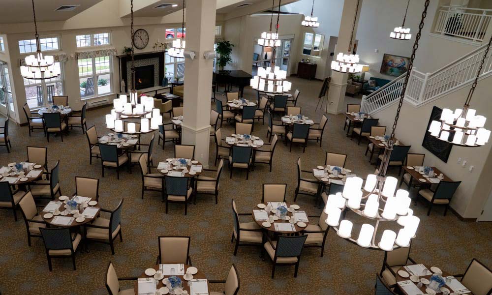 A spacious dining hall with multiple round tables arranged neatly, each set with plates, glasses, and napkins. Large chandeliers hang from the high ceiling, lighting the room brightly. Windows line one wall, and a staircase is visible in the background.