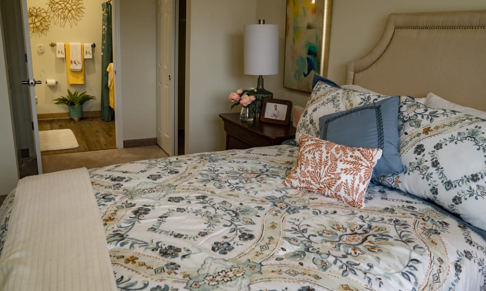 A cozy bedroom with a neatly made bed featuring a floral-patterned comforter and multiple decorative pillows. A nightstand beside the bed holds a lamp, a framed photo, and a vase of flowers. An open door reveals a well-lit, colorful bathroom.