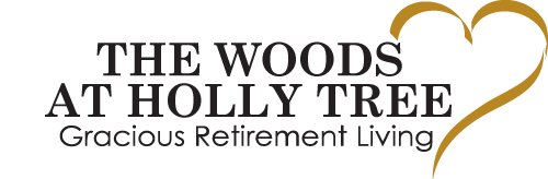 Logo of The Woods at Holly Tree, featuring the text "THE WOODS AT HOLLY TREE: Gracious Retirement Living" in black font. To the right of the text is a gold heart-shaped design.