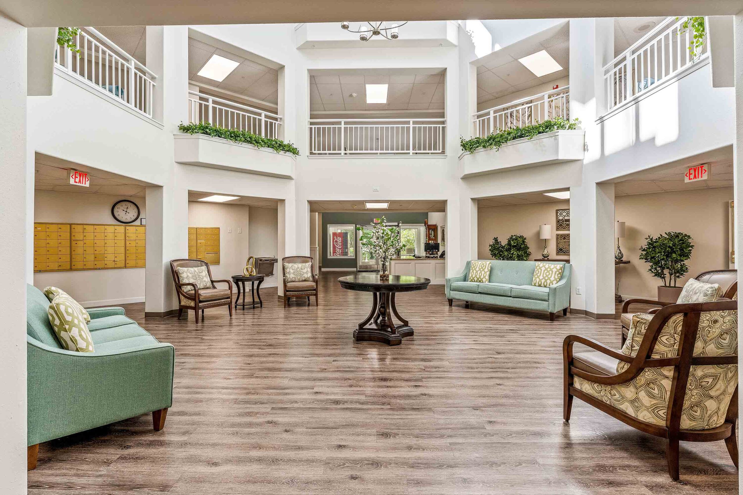 A spacious, well-lit lobby with wooden floors, light green sofas, armchairs, and tables with floral arrangements. Upper-level balcony with plants hanging over the railing, mailboxes on one wall, and multiple exit signs.