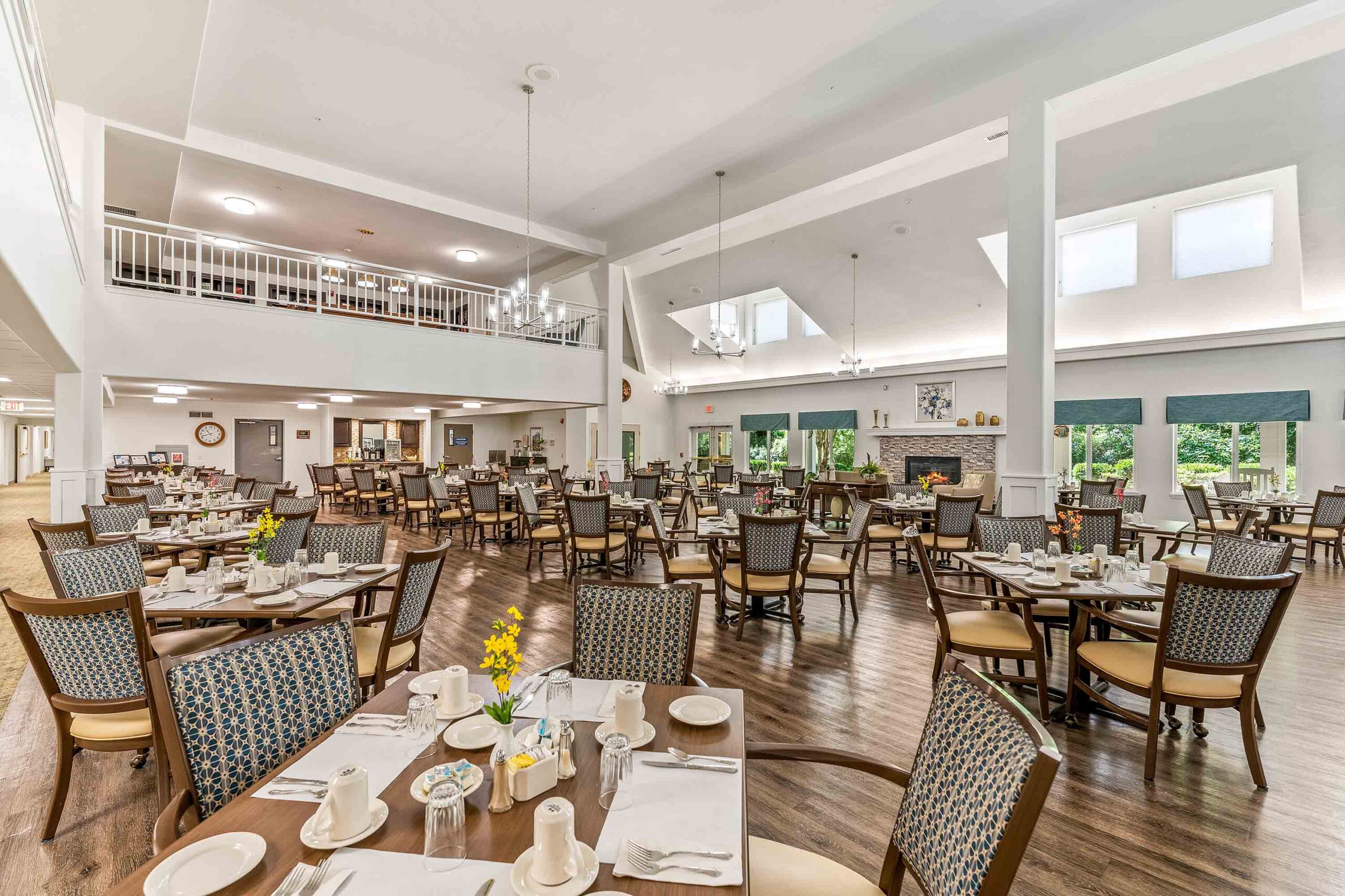 A spacious, well-lit dining room with high ceilings, set with round tables and cushioned chairs. Tables are neatly arranged with white linens, place settings, and small flower vases. Large windows and elegant chandeliers contribute to a welcoming atmosphere.