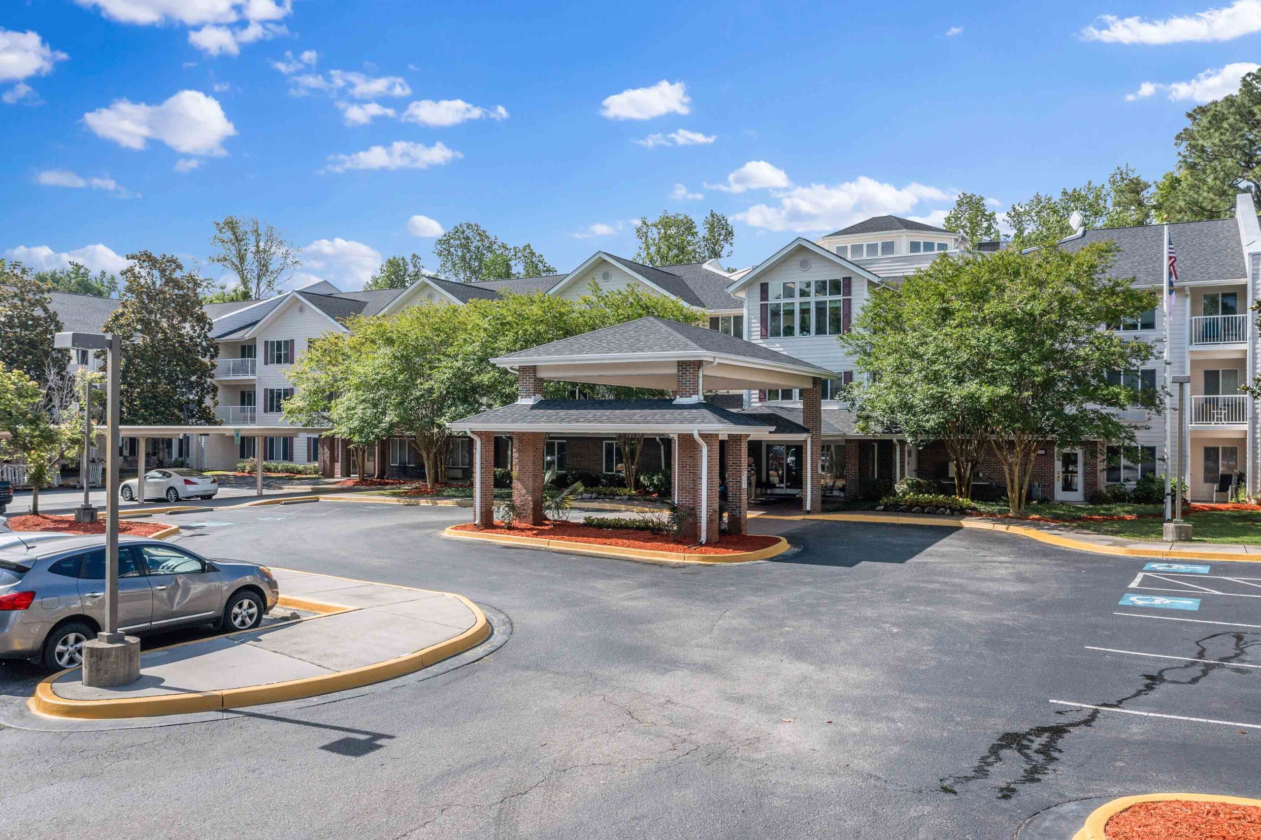 A modern, multi-story residential building with a covered entrance and landscaped surroundings. The parking area in front has several parked cars, and a clear blue sky with scattered clouds is visible in the background.