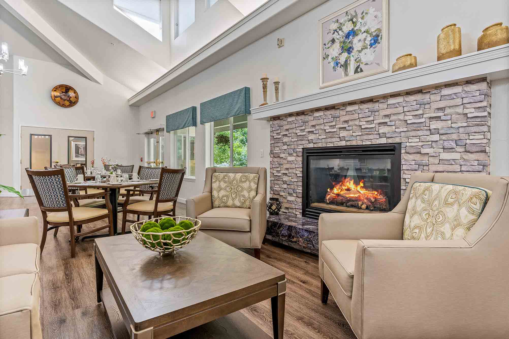 A cozy living room with a modern fireplace surrounded by stone. Two beige armchairs with patterned cushions are placed near the fireplace. A wooden coffee table with a decorative bowl sits in the center. Large windows allow ample natural light into the space.