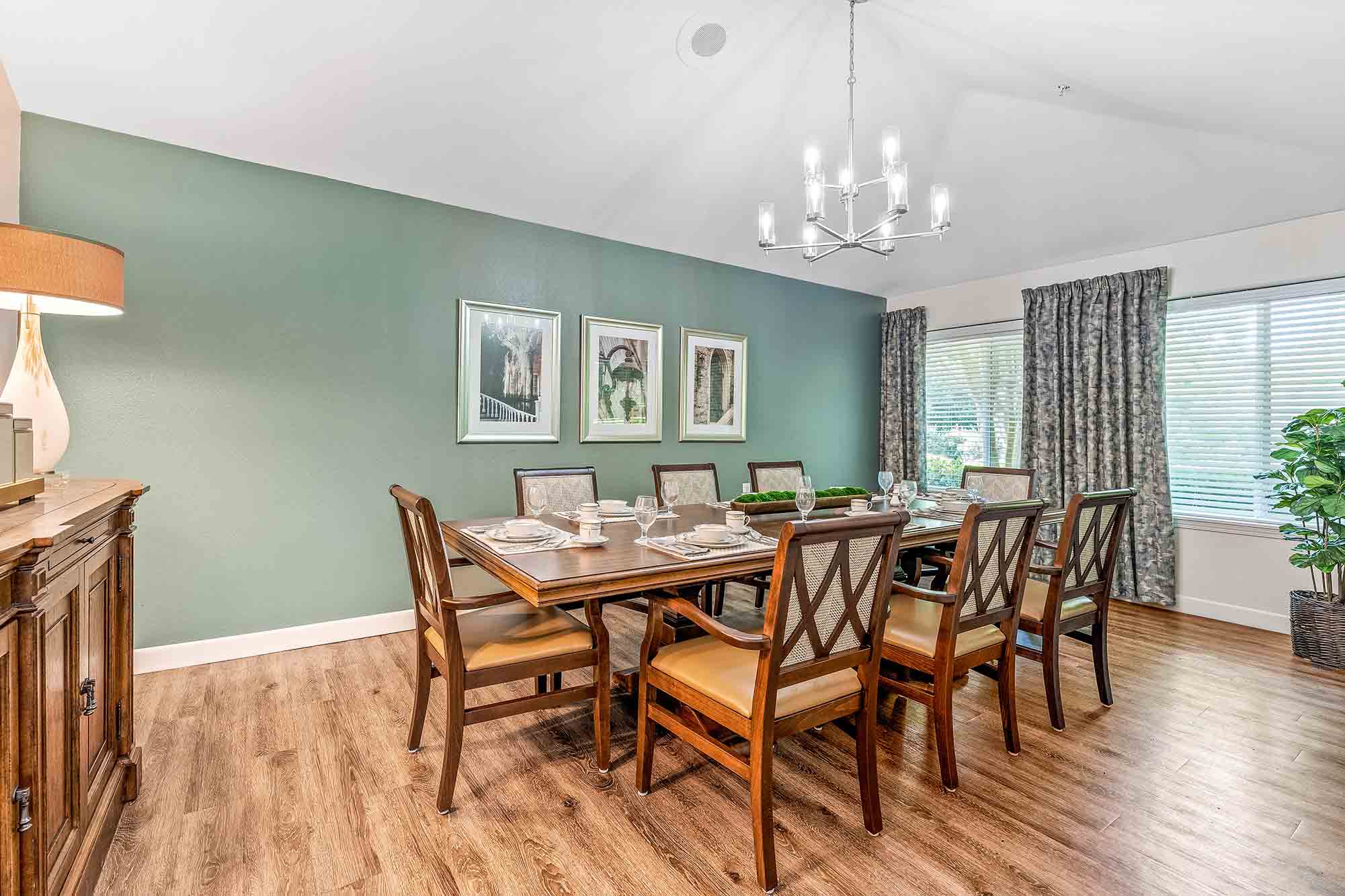 A well-lit dining room with a large wooden table set for six, featuring cushioned chairs. The room has green walls adorned with framed art, a chandelier, a wooden sideboard with a lamp, and large windows with patterned curtains and a potted plant.