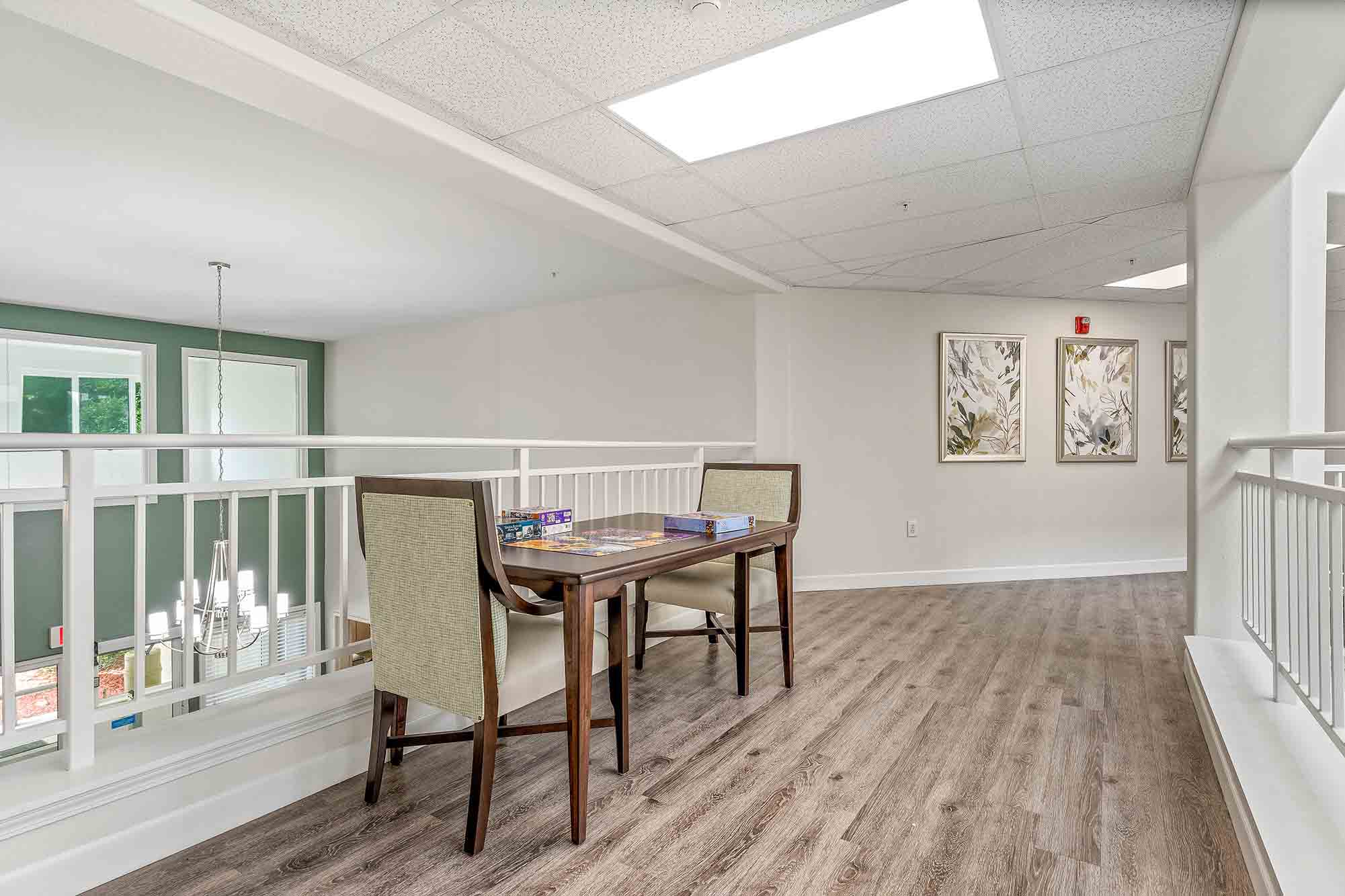 A bright, spacious loft area with wooden flooring, featuring a small table with two upholstered chairs. The table holds puzzles and games. The space is well-lit by overhead lighting, and two decorative framed pictures adorn the wall.