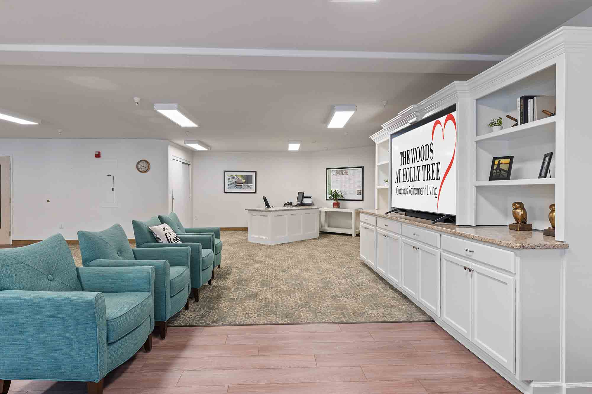 A bright, modern waiting room with teal armchairs, white shelving units, and a large TV displaying 