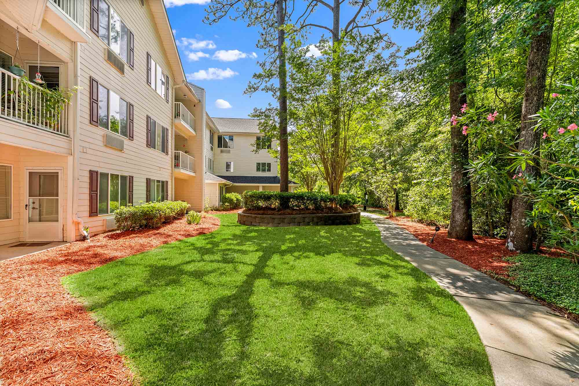 A well-maintained apartment building with light-colored siding is adjacent to a lush, green lawn. Red mulch beds line the sides, and a winding concrete path leads through the grassy area. Tall trees and various plants provide a natural, serene backdrop.