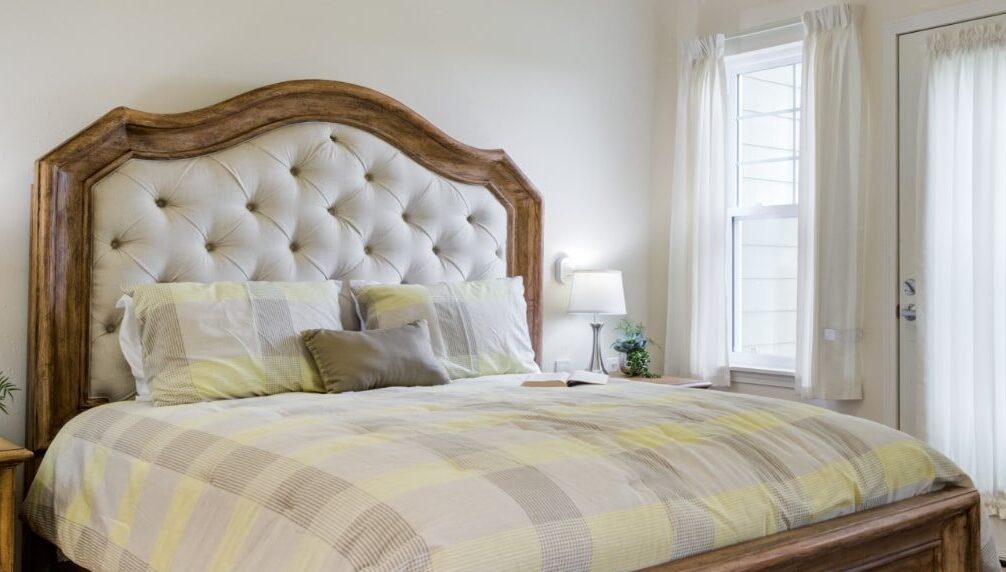 A cozy bedroom with a large bed featuring a tufted headboard made of light-colored fabric and wooden frame. The bed is adorned with plaid bedding in shades of yellow, beige, and gray, along with multiple pillows. A small nightstand with a lamp and a window are beside the bed.