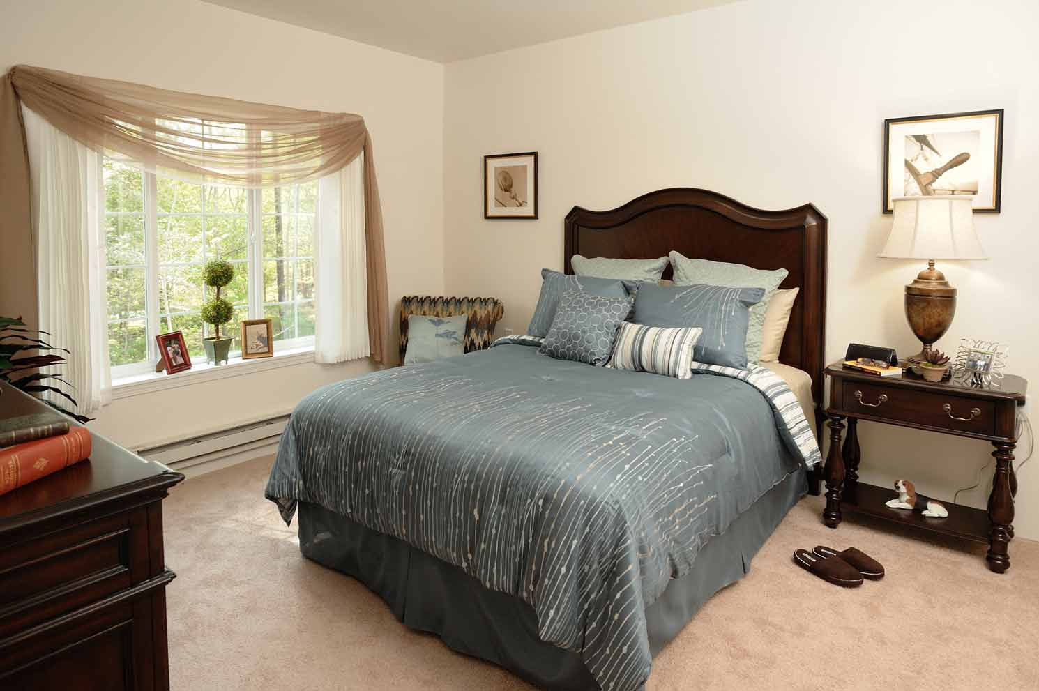 A cozy bedroom featuring a large bed with blue and grey bedding, flanked by two wooden nightstands with lamps. A bay window with sheer curtains lets in natural light. The room is decorated with framed pictures, a potted plant, and a pair of slippers on the floor.