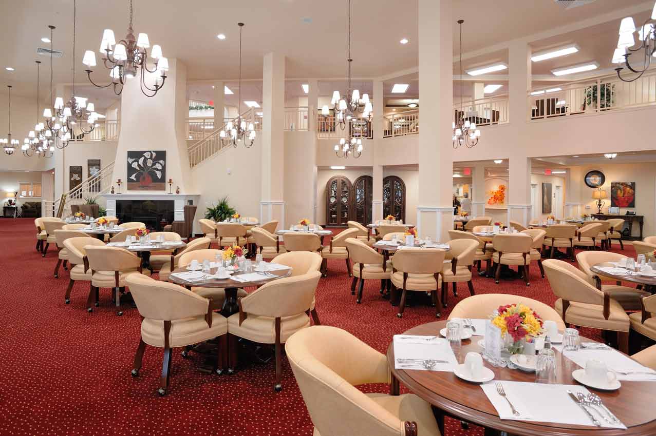 A spacious, elegantly decorated dining hall with red carpet and beige chairs arranged around tables set with white tablecloths, dishes, and silverware. Several chandeliers hang from the high ceiling, and a staircase and second-floor balcony are visible in the background.