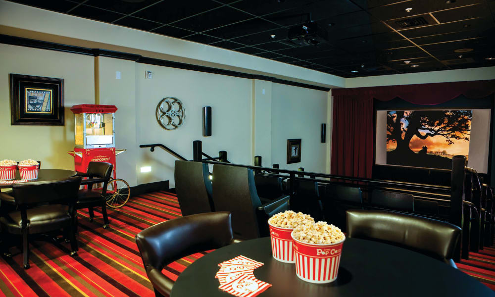 A cozy home theater room with black leather chairs and red-striped carpet. A small table with popcorn buckets and cards sits in the foreground. A popcorn machine is against the wall, and a projection screen shows a movie scene with a tree at sunset.