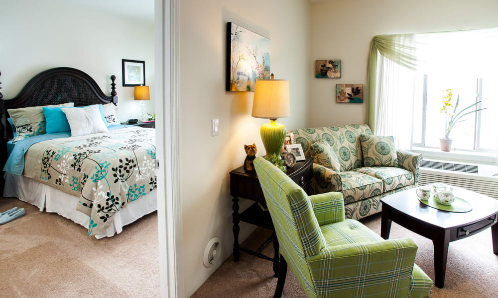 A cozy apartment's interior featuring an open layout. On the left, a bedroom with a dark wooden bed, teal and white bedding, and floral decorations. On the right, a small living area with green plaid chairs, a floral couch, and a lamp on a side table. A window lets in natural light.