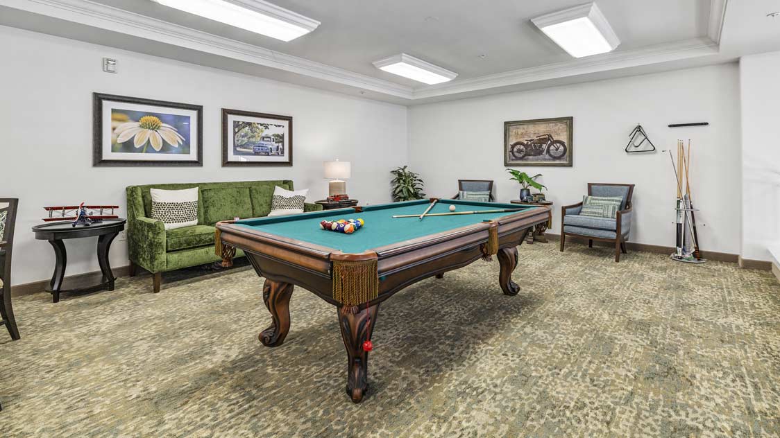 A room with a pool table in the center, surrounded by green and gray furniture. Art pieces and photographs adorn the walls. A coat rack and pool cues are in the corner. The floor is carpeted, and the area is well-lit with ceiling lights.