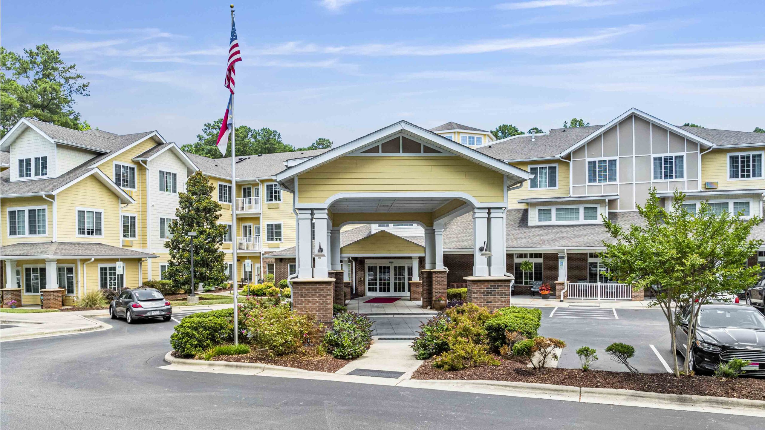 A well-maintained assisted living facility with multiple stories and a large entrance covered by a yellow canopy. The facility is surrounded by lush greenery and flowerbeds. An American flag and a state flag are flying on flagpoles. Several cars are parked outside.