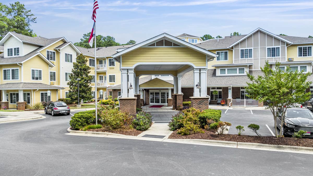 A modern, multi-storied residential building with white and yellow exterior, featuring a covered entrance and landscaped surroundings. A flagpole with a flag stands near the entrance, and several parked cars are visible in the foreground.