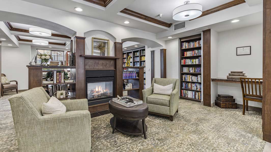 A cozy library room with light green armchairs around a coffee table, a central fireplace, floor-to-ceiling bookshelves, and a wooden desk with a chair in the corner. The room has a beige carpet, wood-beamed ceiling, and pendant lights.