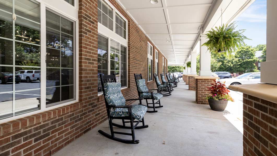 A covered porch with brick walls features a row of black wooden rocking chairs with patterned cushions. Potted plants and hanging ferns decorate the space, and cars are parked in the background. Large windows provide a view inside the building.