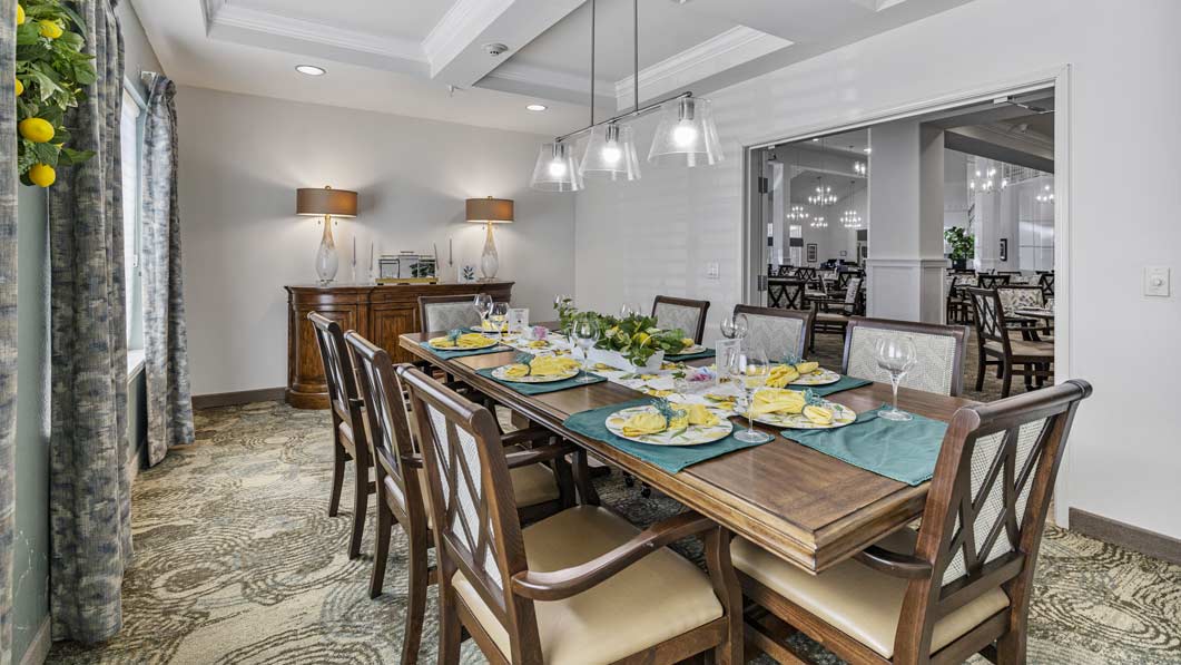 A dining room with a wooden table set for eight people, featuring teal placemats and yellow napkins. The room has cushioned chairs, a sideboard with two lamps, and an open doorway leading to another room with additional dining tables. Pendant lights hang above the table.