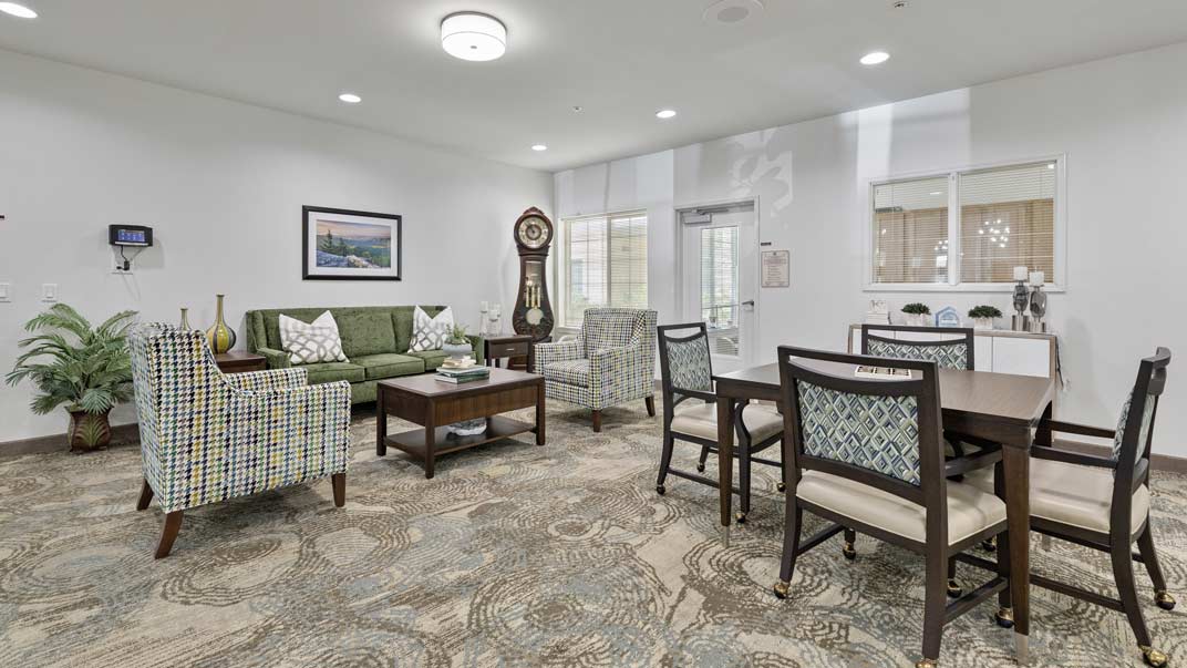 A spacious and well-lit living room with modern furniture. There is a green sofa, plaid-patterned armchairs, a wooden clock, and a glass coffee table. A large rug covers the floor, and there are potted plants adding a touch of greenery to the space.