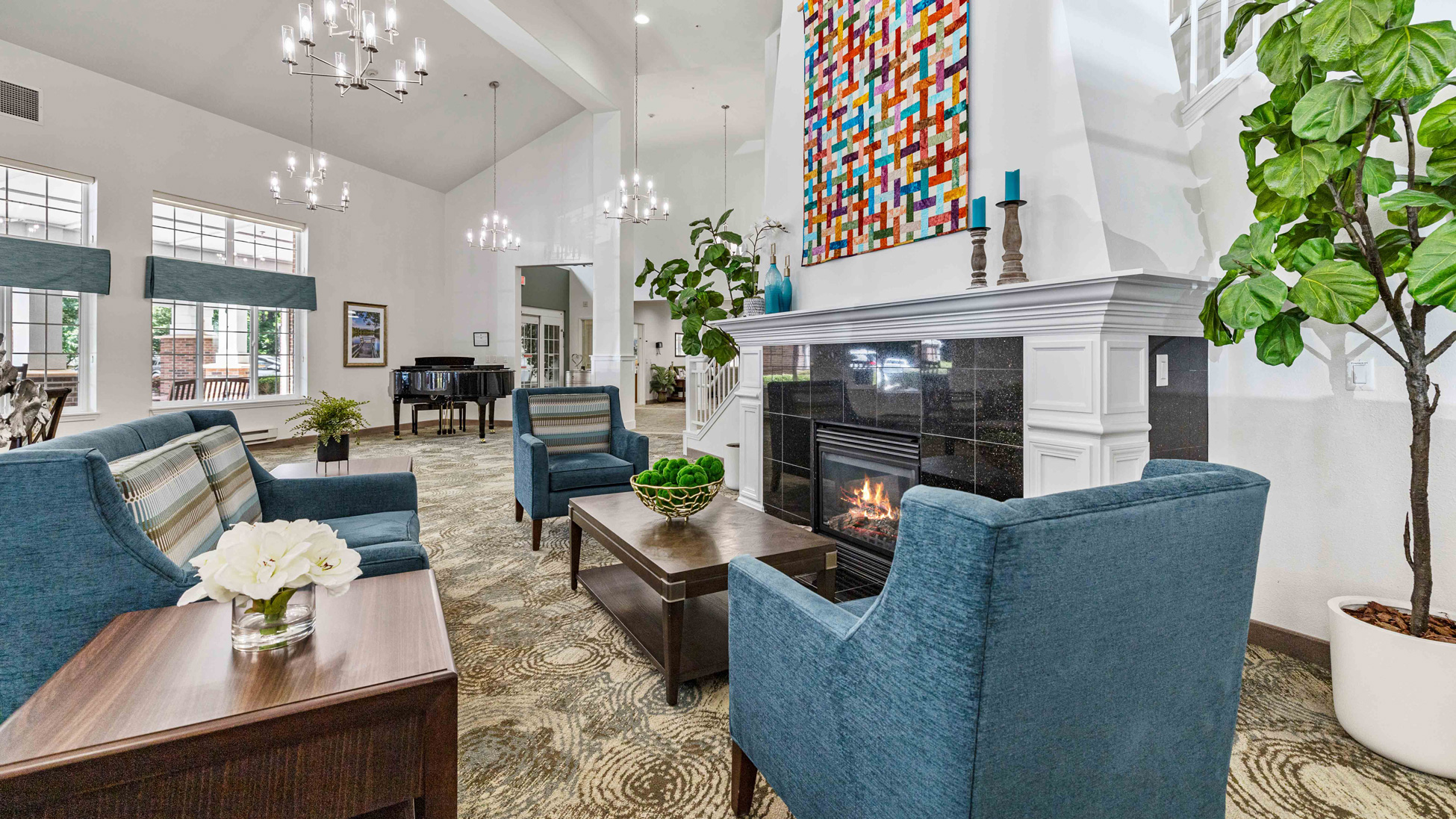 A modern, bright living room with blue armchairs, a coffee table, a fireplace, a colorful abstract painting above the mantel, large windows, plants, and a piano in the background.