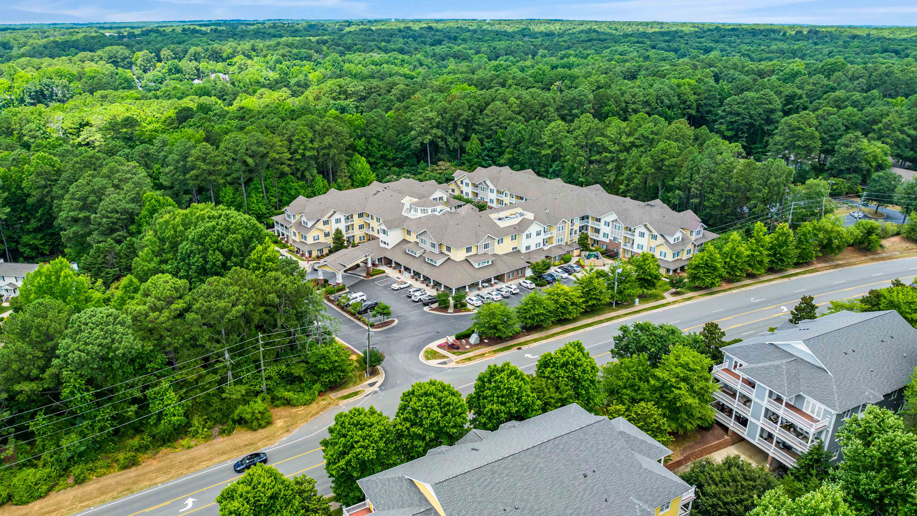 Aerial view of a large residential complex surrounded by dense green trees, with parking lots and cars visible. The complex is adjacent to a main road, with other buildings and forested areas nearby.