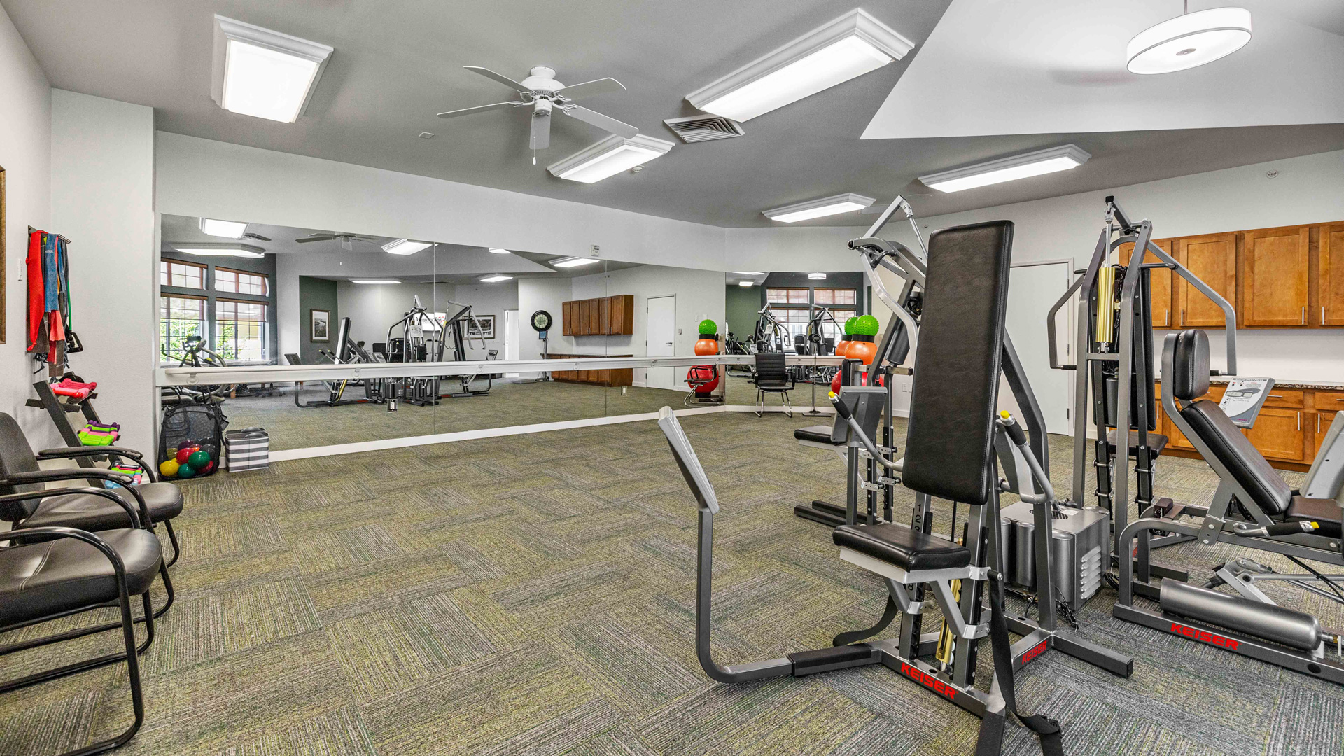 A spacious gym room with various exercise machines, free weights, resistance bands, exercise balls, and several chairs. Large mirrors cover one wall, and windows let in natural light. Cabinets and a sink are in the back corner.