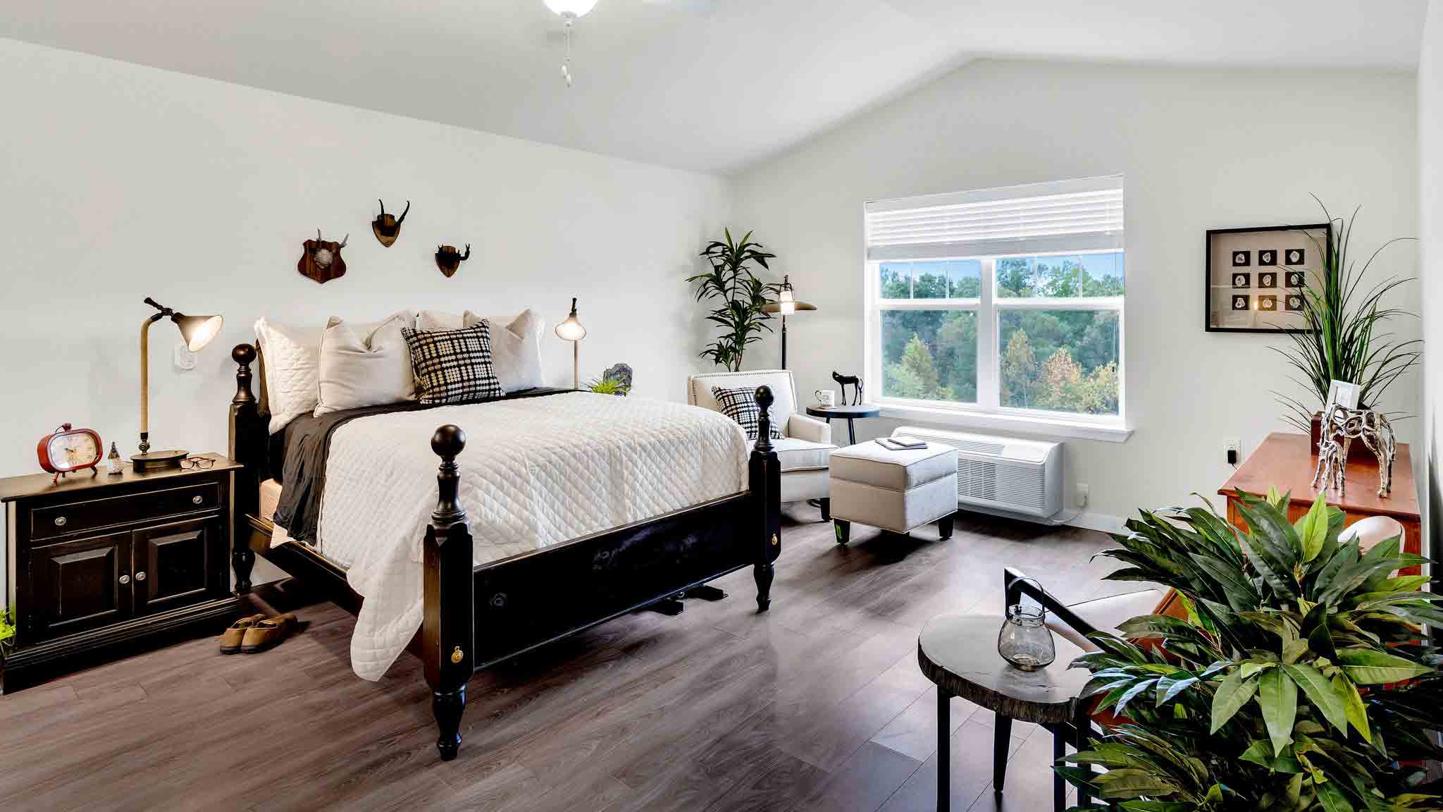 A bright bedroom with a black bed frame, white bedding, two black nightstands, a beige armchair by a large window, wooden floor, and several plants. Decor includes wall art and mounted antlers.