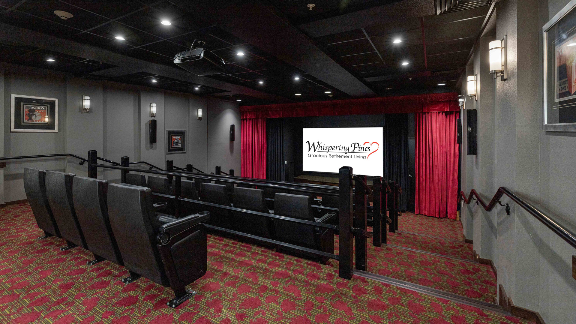 A small movie theater with tiered seating, red patterned carpet, framed pictures on the walls, and a large screen displaying "Whispering Pines Gracious Retirement Living.