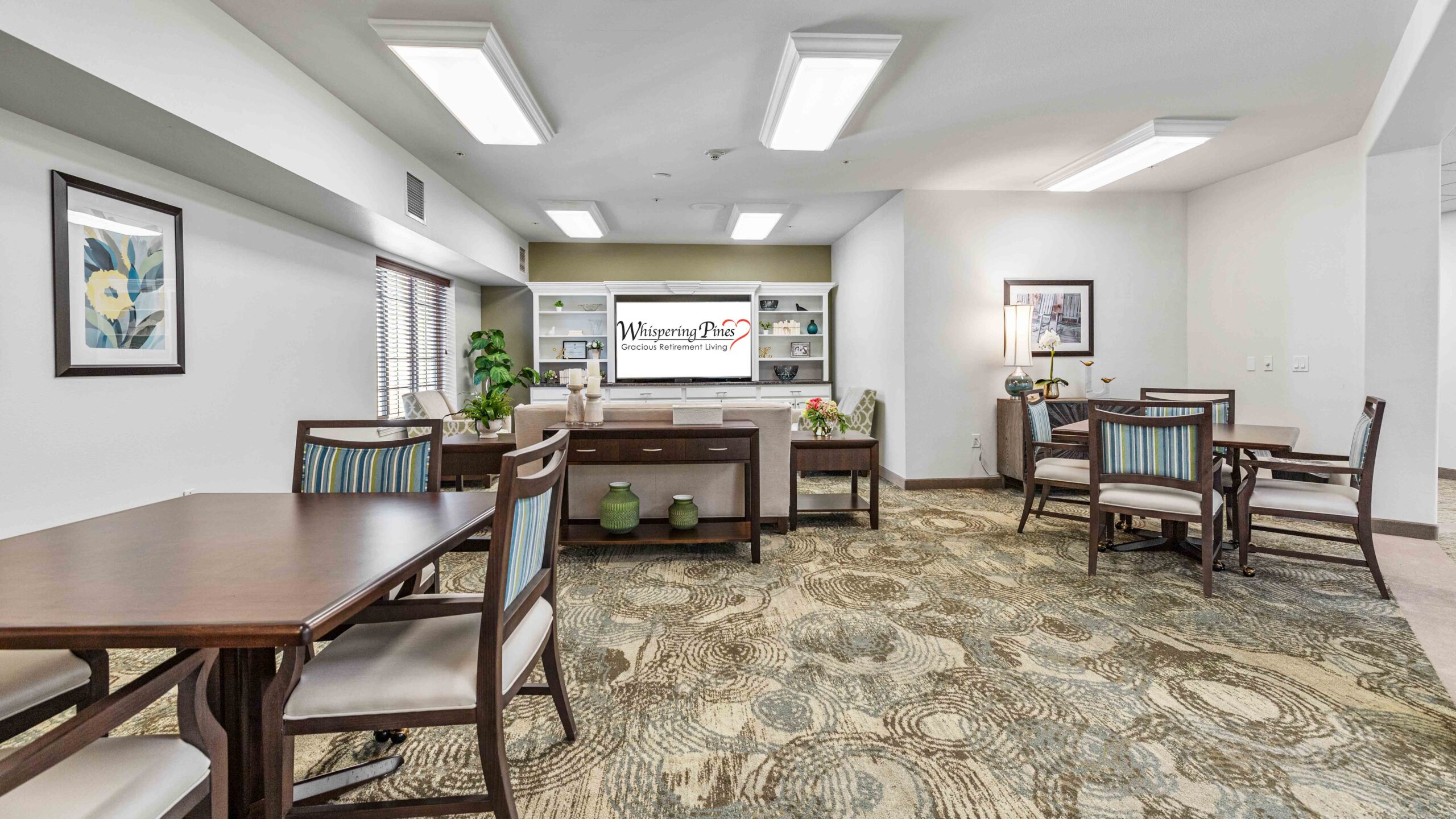 A spacious, well-lit common room with wooden tables and chairs, cushioned seating, artwork on the walls, shelves with decorative items, and patterned carpet. Large windows let in natural light.