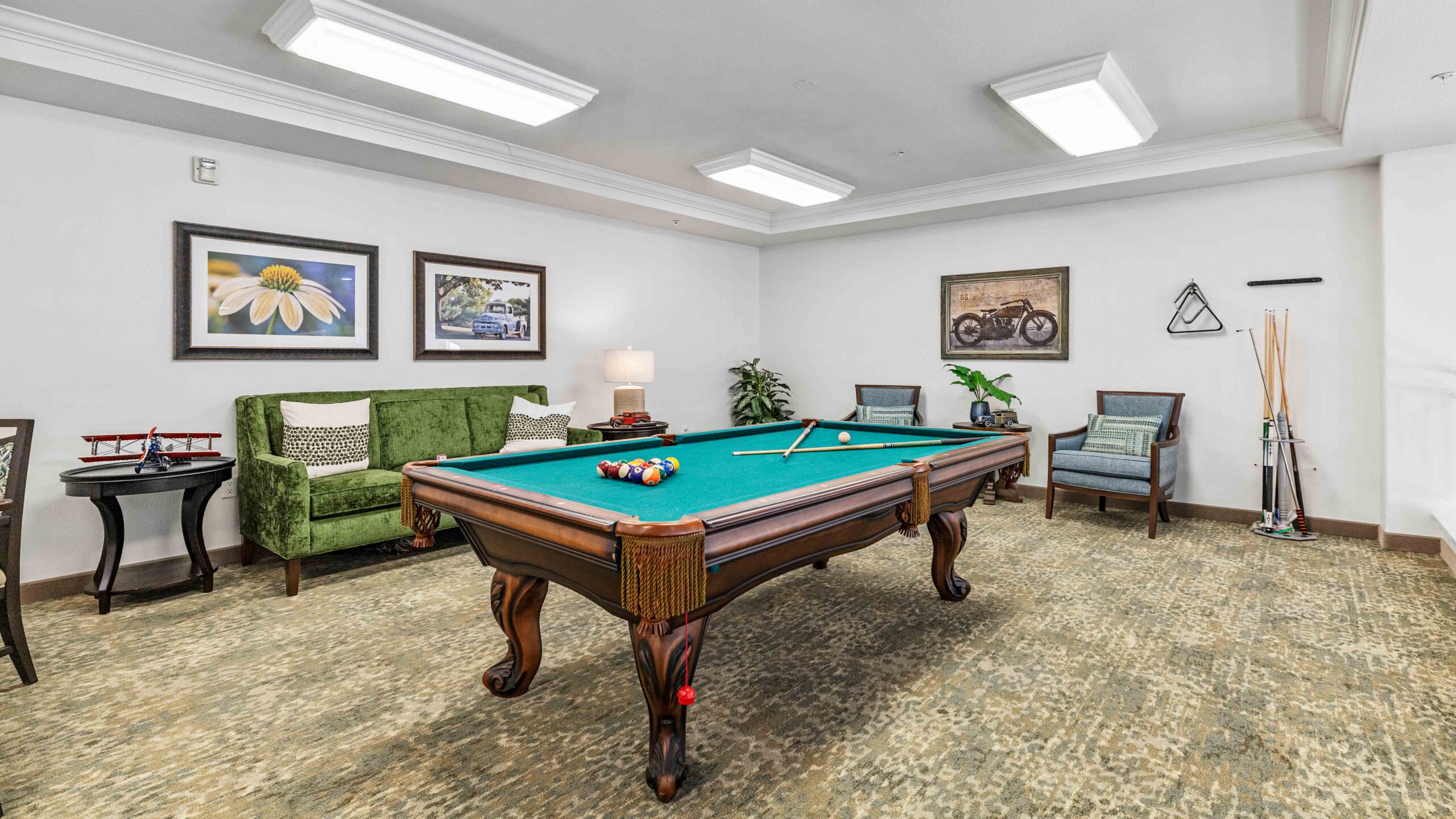 A well-lit room with a pool table in the center, balls and cues on top, green sofa, two armchairs, framed artwork on the walls, and a lamp on a side table. Pool cues are stored in a corner rack.