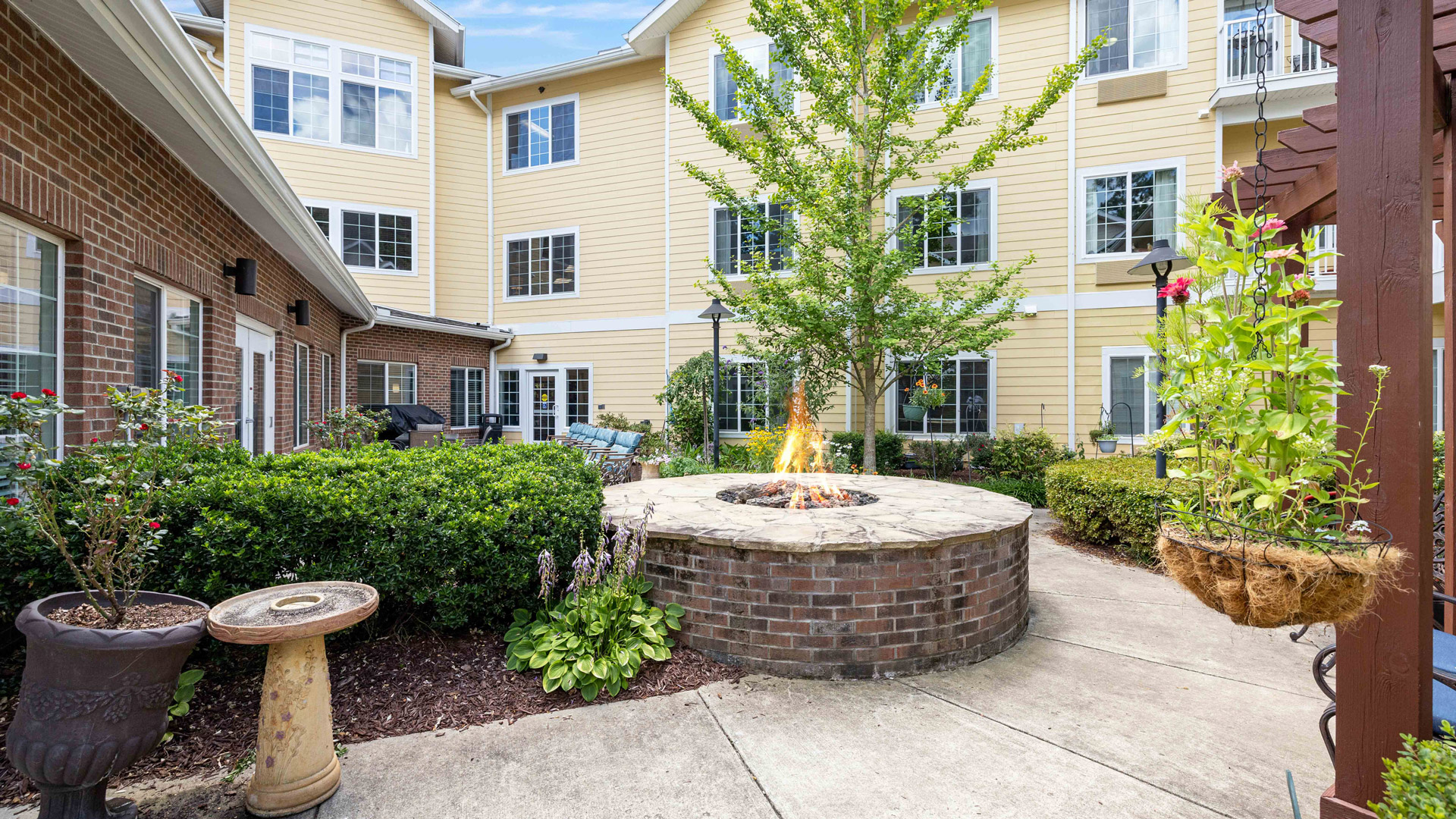 A courtyard with a circular stone fire pit, green shrubs, potted plants, and pathways, surrounded by a yellow and brick multi-story building with many windows.