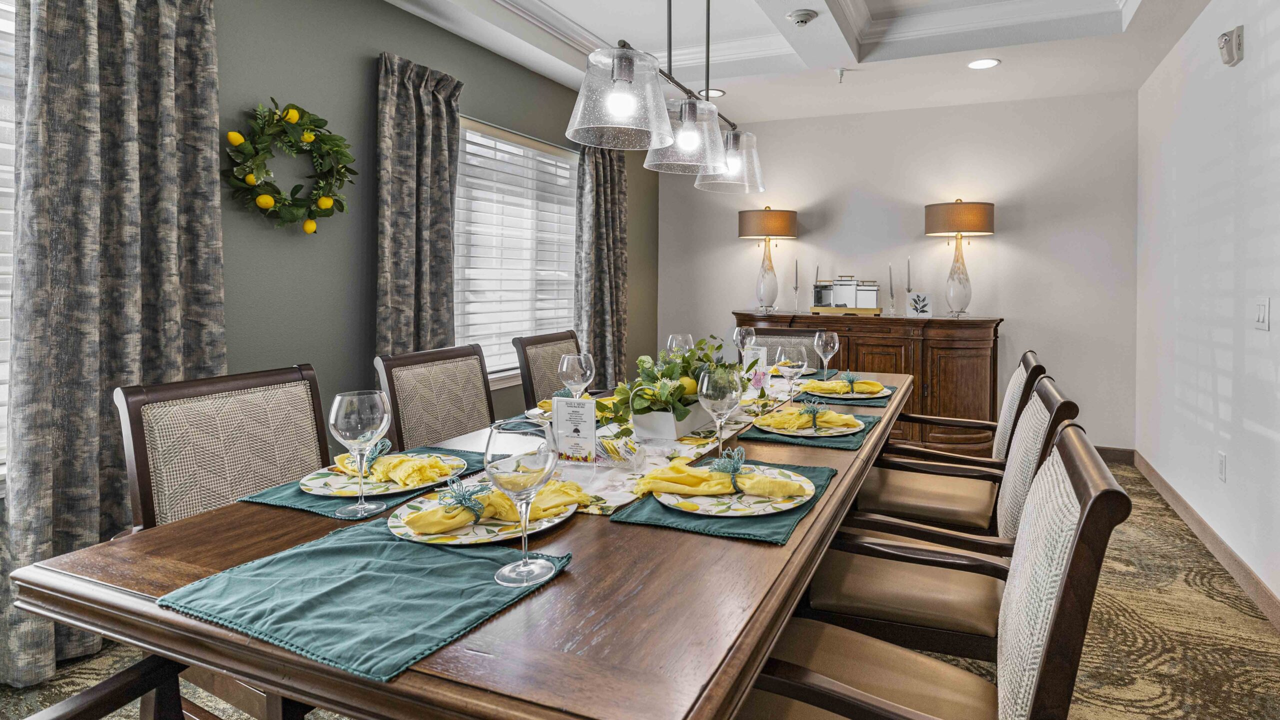 Elegant dining room with a long wooden table set for six, featuring yellow napkins, wine glasses, and floral centerpiece. A sideboard with lamps and candles is against the back wall, and a lemon wreath hangs on a green accent wall.