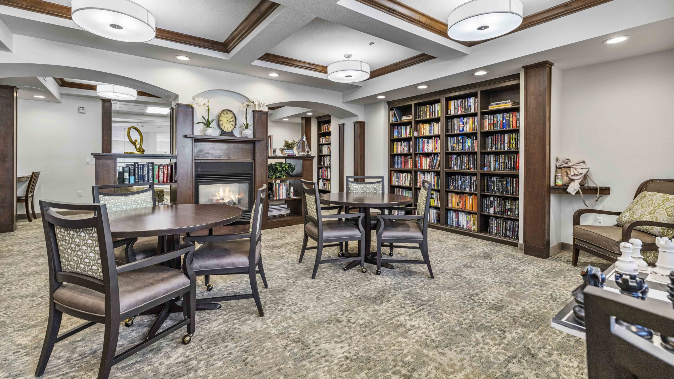 A cozy library room with bookshelves lining the wall, round tables with chairs, a lit fireplace, and modern ceiling lights. There are armchairs and a chess set, creating a warm and inviting atmosphere.