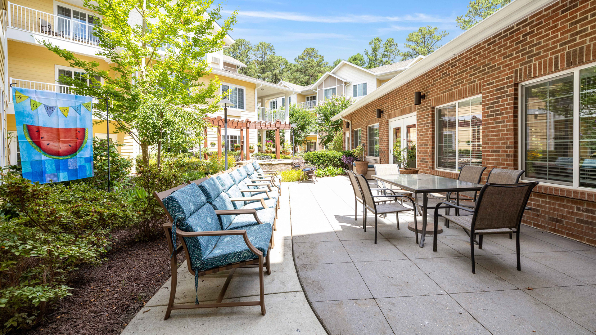 Sunny outdoor patio with cushioned chairs lined along a wall, a glass-top table with chairs, brick and yellow buildings, green plants, and a colorful towel featuring a watermelon design hanging nearby.